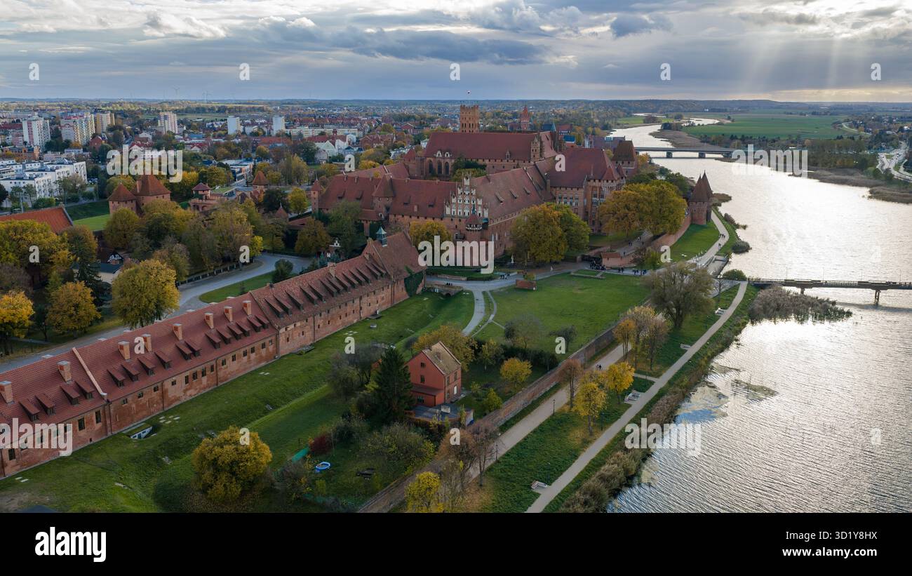 Die Drohnenansicht zeigt die Burg Malbork in Malbork, Polen, mit Türmen, Mauern, Innenhöfen, und eine Fußgängerbrücke am Fluss am späten Nachmittag Stockfoto