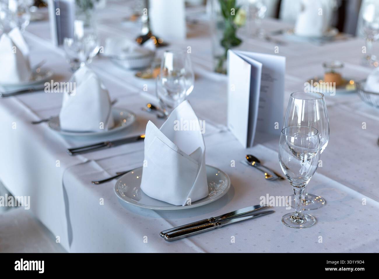 An einem lebhaften Tisch im Restaurant finden Sie ordentlich angeordnete Teller mit gefalteten Servietten, glänzende Gläser und Menüs. Die sanfte Beleuchtung sorgt für ein angenehmes Ambiente Stockfoto