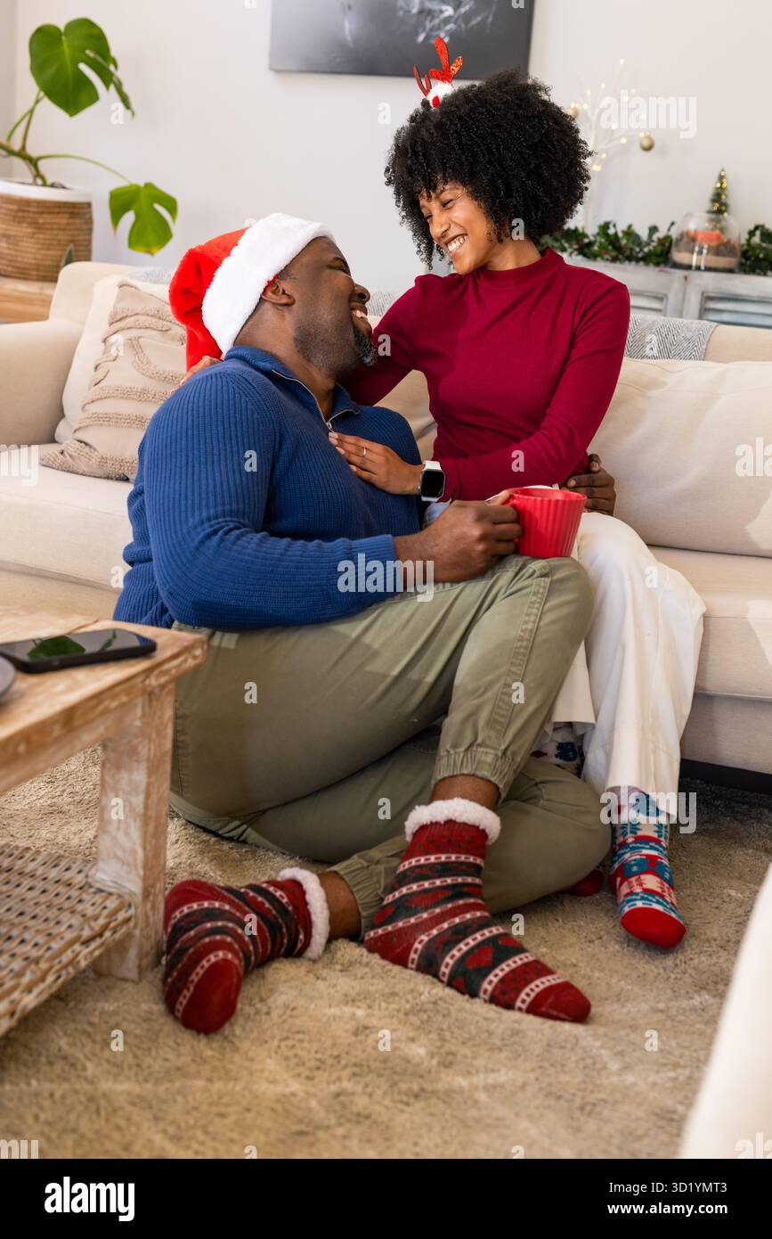 Afroamerikanisches Paar mit festlicher Kopfbedeckung, das im Wohnzimmer sitzt und einen roten Becher lacht Stockfoto