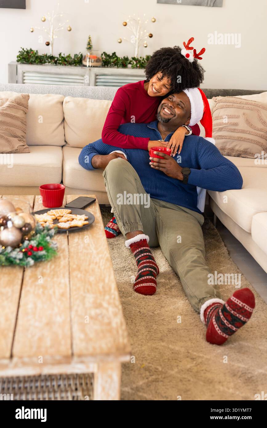 Afroamerikanisches Paar mit Pullover, die zu Hause Feiertage feiern und rote Tassen und Kekse halten Stockfoto