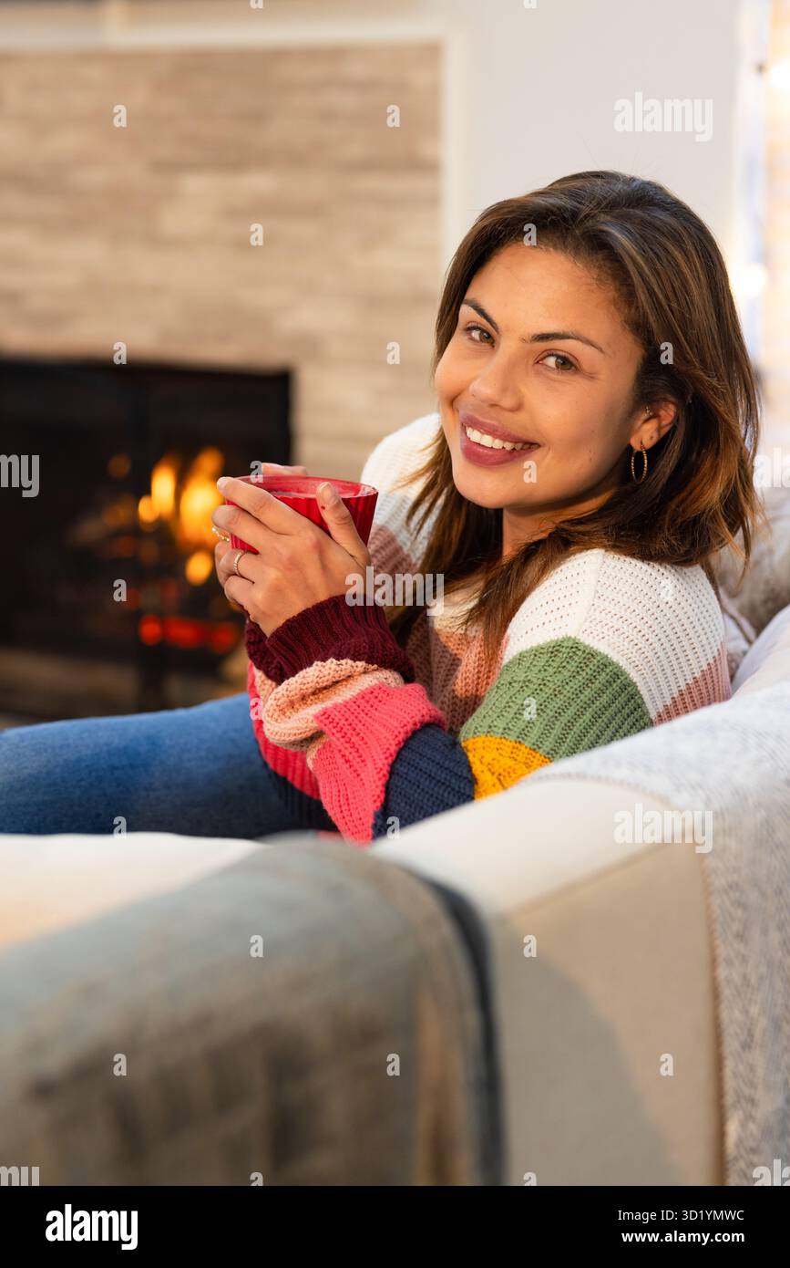 MittelErwachsene Frau, die zu Hause sitzt und einen roten Becher hält, während sie sich unter einer Decke am Kamin kuschelt Stockfoto