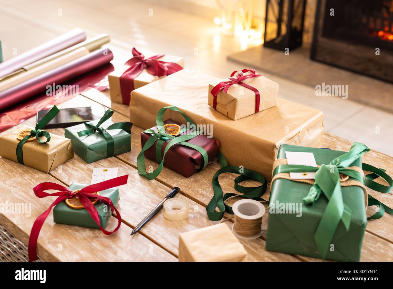 Gewickelte Geschenke, die auf einer Holzplatte in der Nähe des glühenden Kamins liegen, mit Bändern, getrockneten orangefarbenen Tags Stockfoto