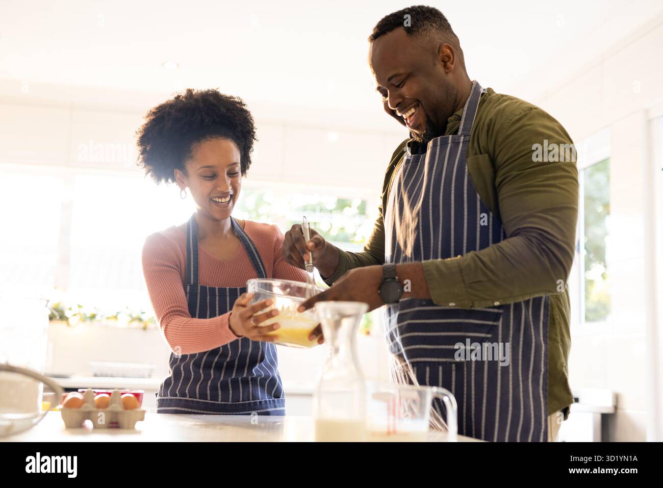 Afroamerikanisches Paar, das gestreifte Schürzen trägt und Eier in einer Glasschale mit Schneebesen in der Küche mischt Stockfoto