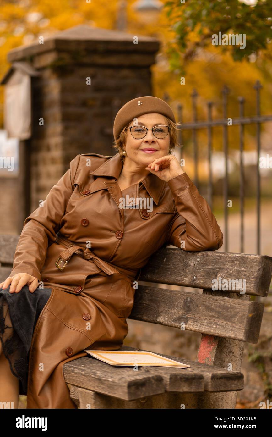 Eine stilvolle Frau genießt die Ruhe eines Parks, während sie auf einer Holzbank sitzt. Die Szene ist mit leuchtenden Herbstfarben wie Orange und Gelb gefüllt Stockfoto