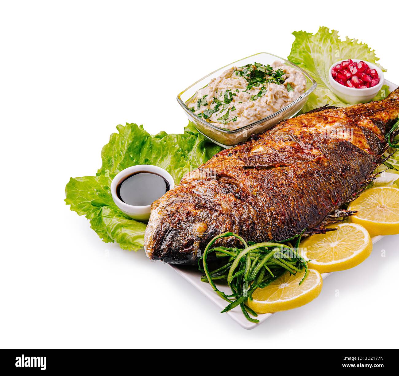 Gegrillter ganzer Fisch serviert mit frischen Kräutern und Beilagen auf einer weißen Platte, garniert mit Zitronenscheiben und Gemüse Stockfoto