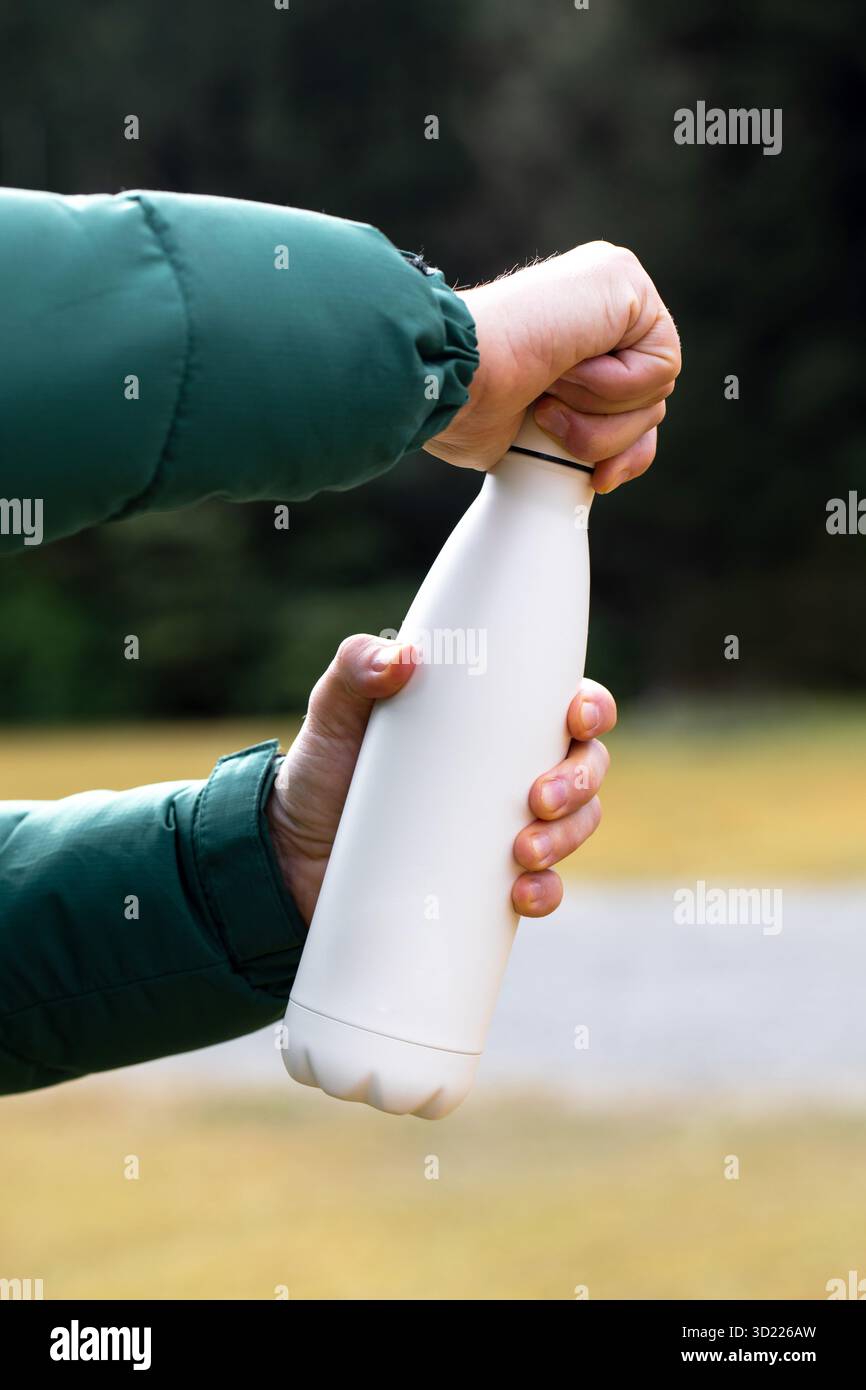 Man Hand hält Modell-Thermos gegen verschwommene Büsche aus der Nähe. Touristenshows Wasserflasche mit Kopierraum im Freien. Mehrwegkolben Stockfoto