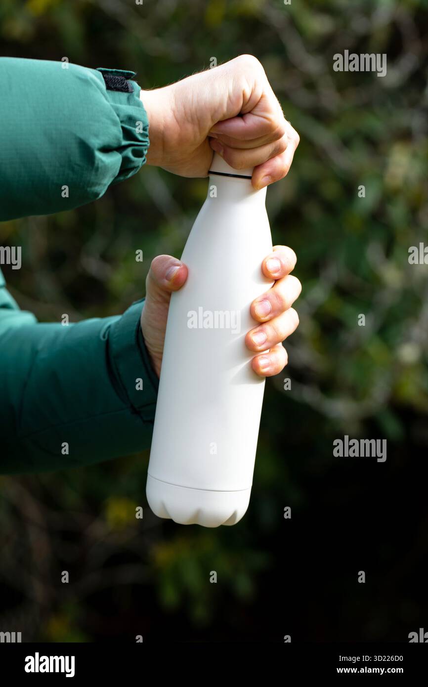 Man Hand hält Modell-Thermos gegen verschwommene Büsche aus der Nähe. Touristenshows Wasserflasche mit Kopierraum im Freien. Mehrwegkolben Stockfoto