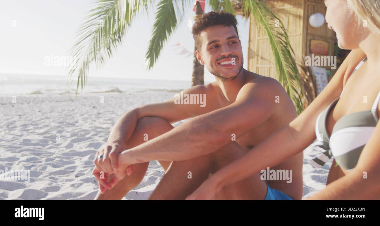 Lächelndes Paar, das auf Sand unter Palmen von Meereswellen sitzt, mit Strohdach-Hütte und Papierlaternen Stockfoto