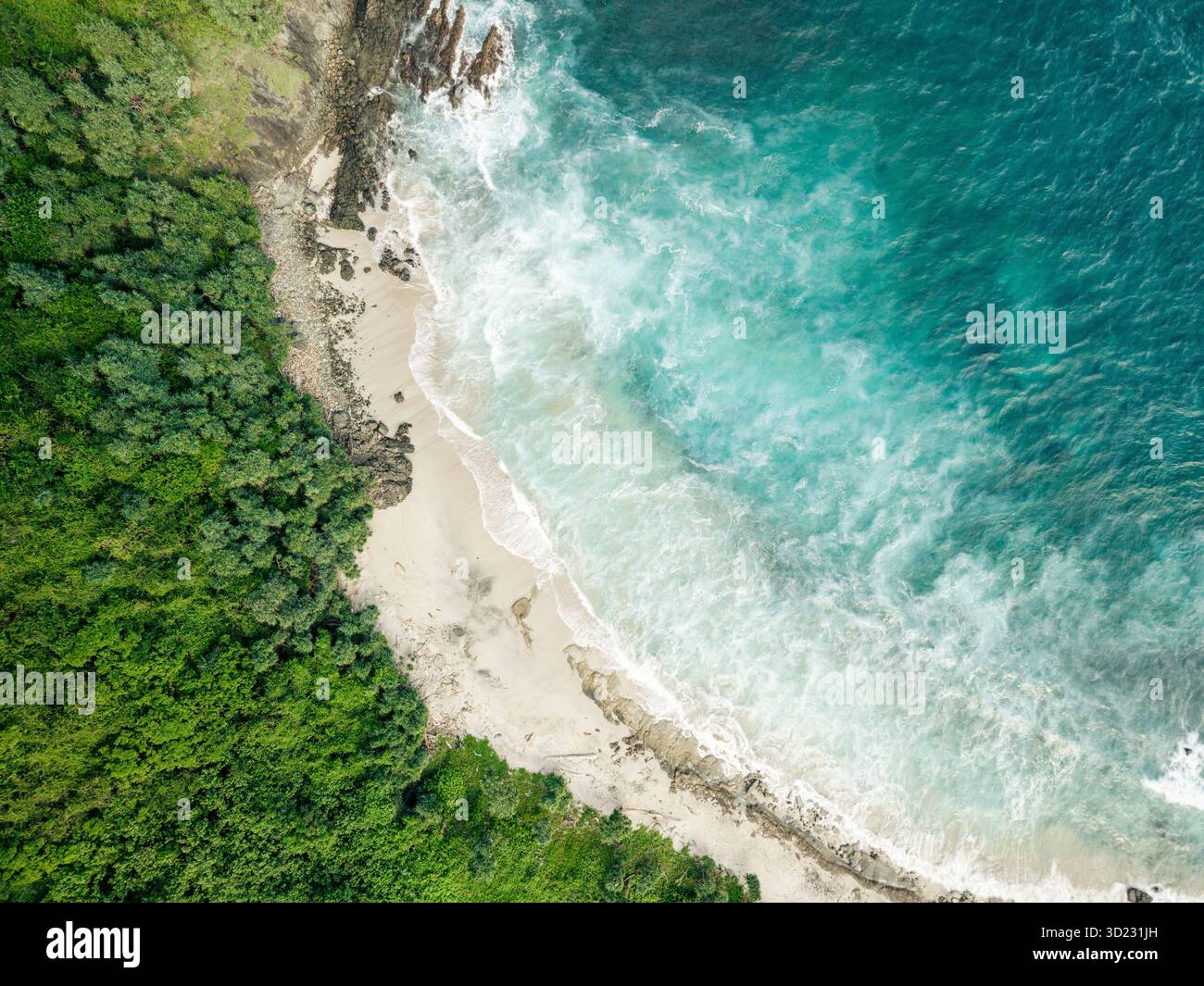 Aus der Vogelperspektive auf eine üppige grüne Küste, die auf die pulsierenden blauen Wellen des Ozeans an einem Sandstrand trifft. Lombok, Mawi, Indonesien Stockfoto