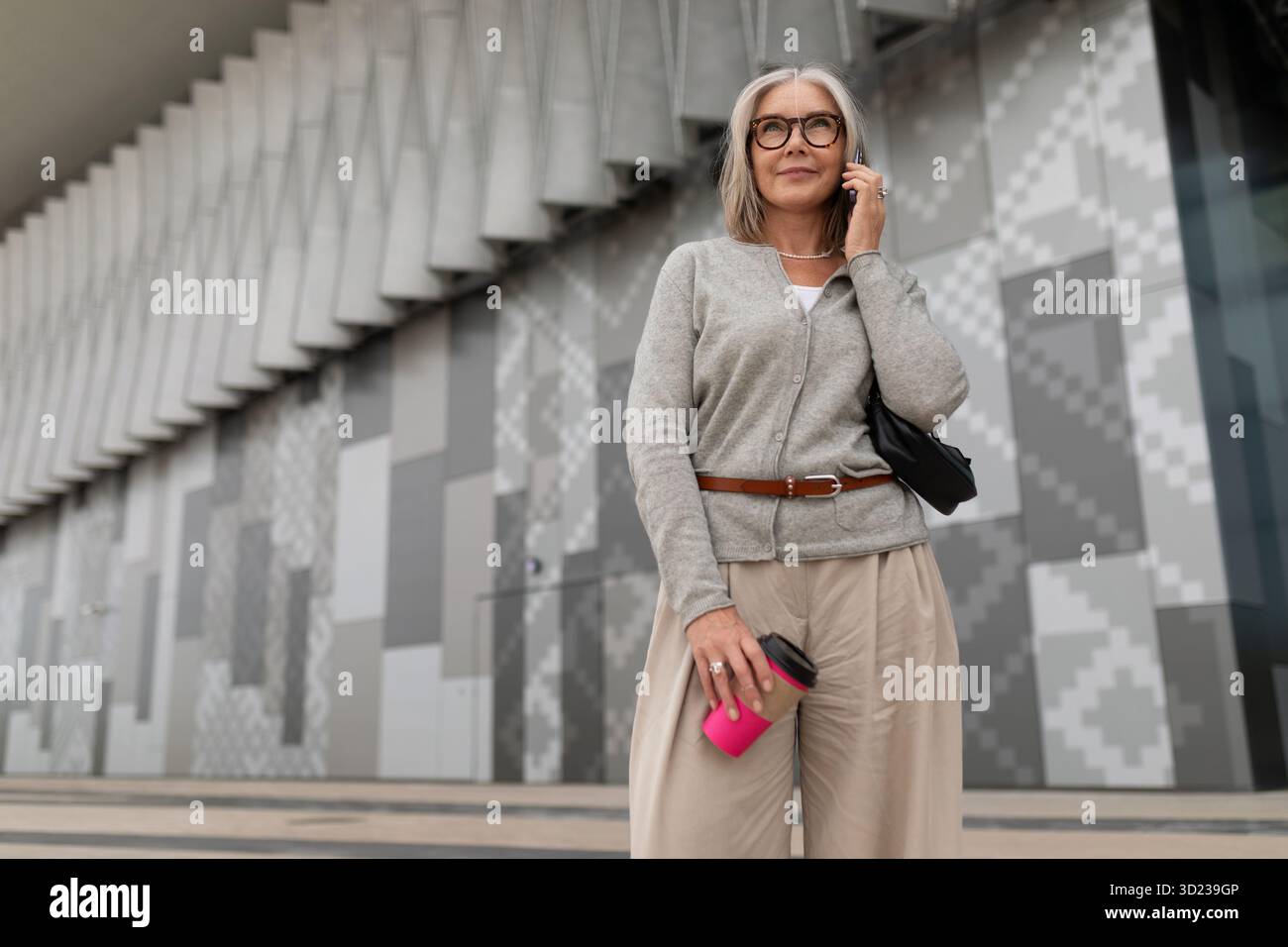 Frau in legeren Kleidern steht draußen mit Kaffee, während sie ein Smartphone in einer modernen urbanen Umgebung benutzt Stockfoto