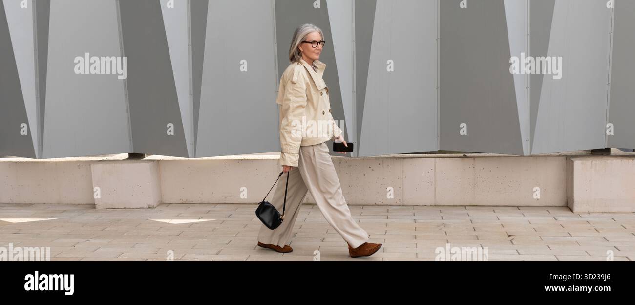 Elegante ältere Frau geht selbstbewusst entlang eines stilvollen urbanen Weges neben modernem architektonischen Design Stockfoto