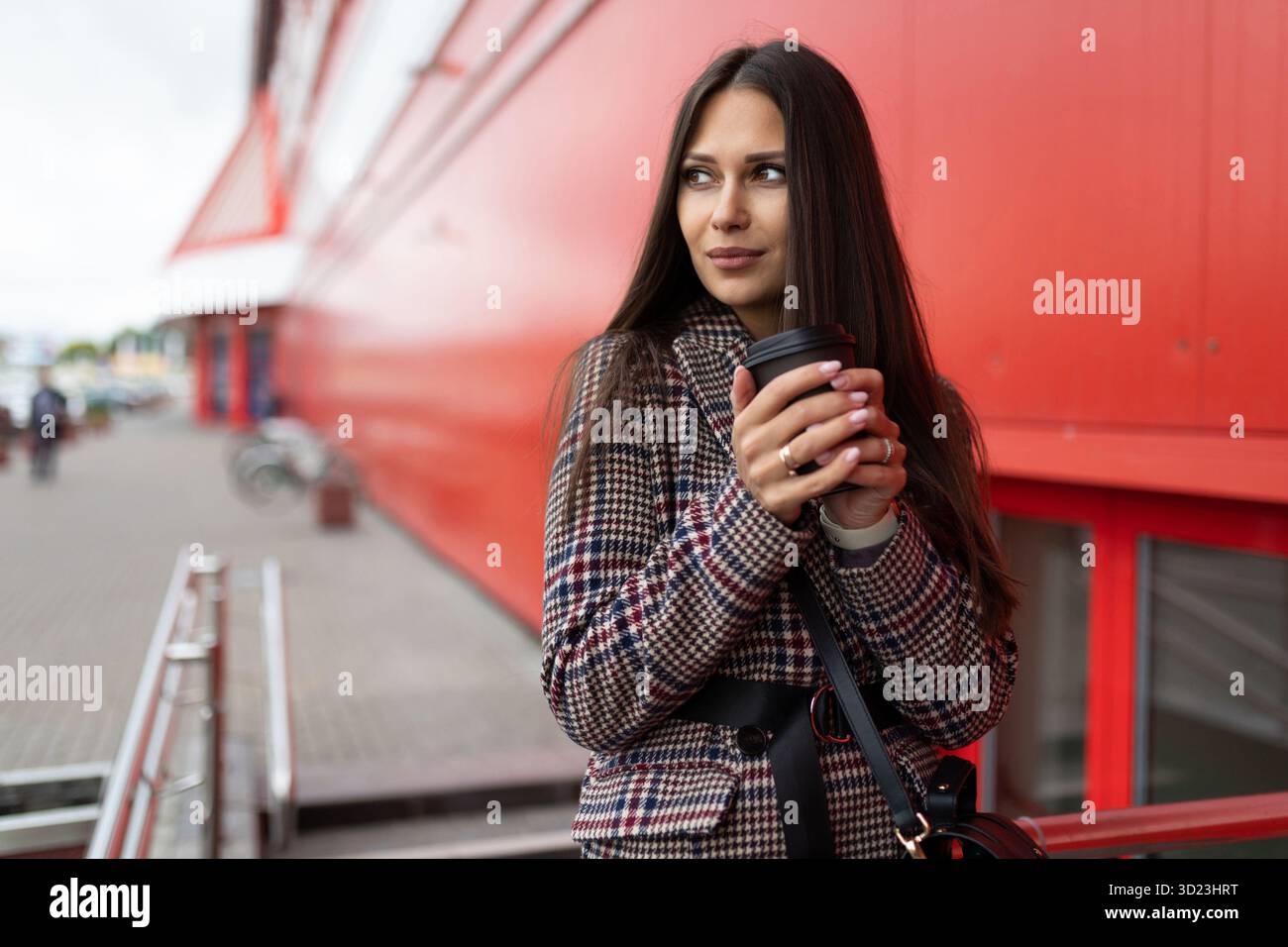Eine junge Frau mit einer Tasse Tee in der Hand wärmt ihre Hände mit einem Lächeln, der in die Ferne neben dem Einkaufszentrum blickt Stockfoto