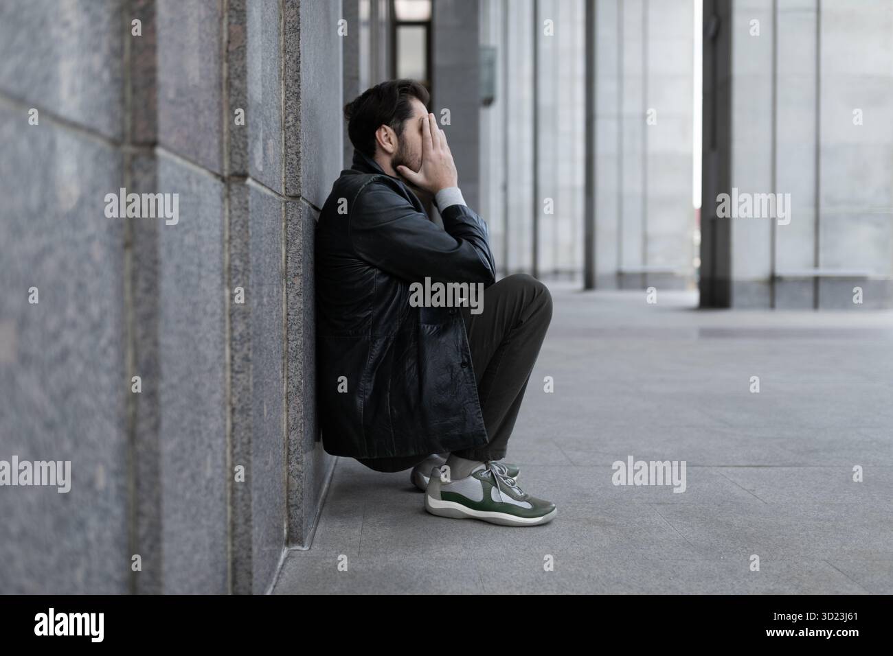 Ein Mann in einem deprimierten Zustand sitzt zurück an der Wand und bedeckt seine Hände mit seinen Handflächen Stockfoto
