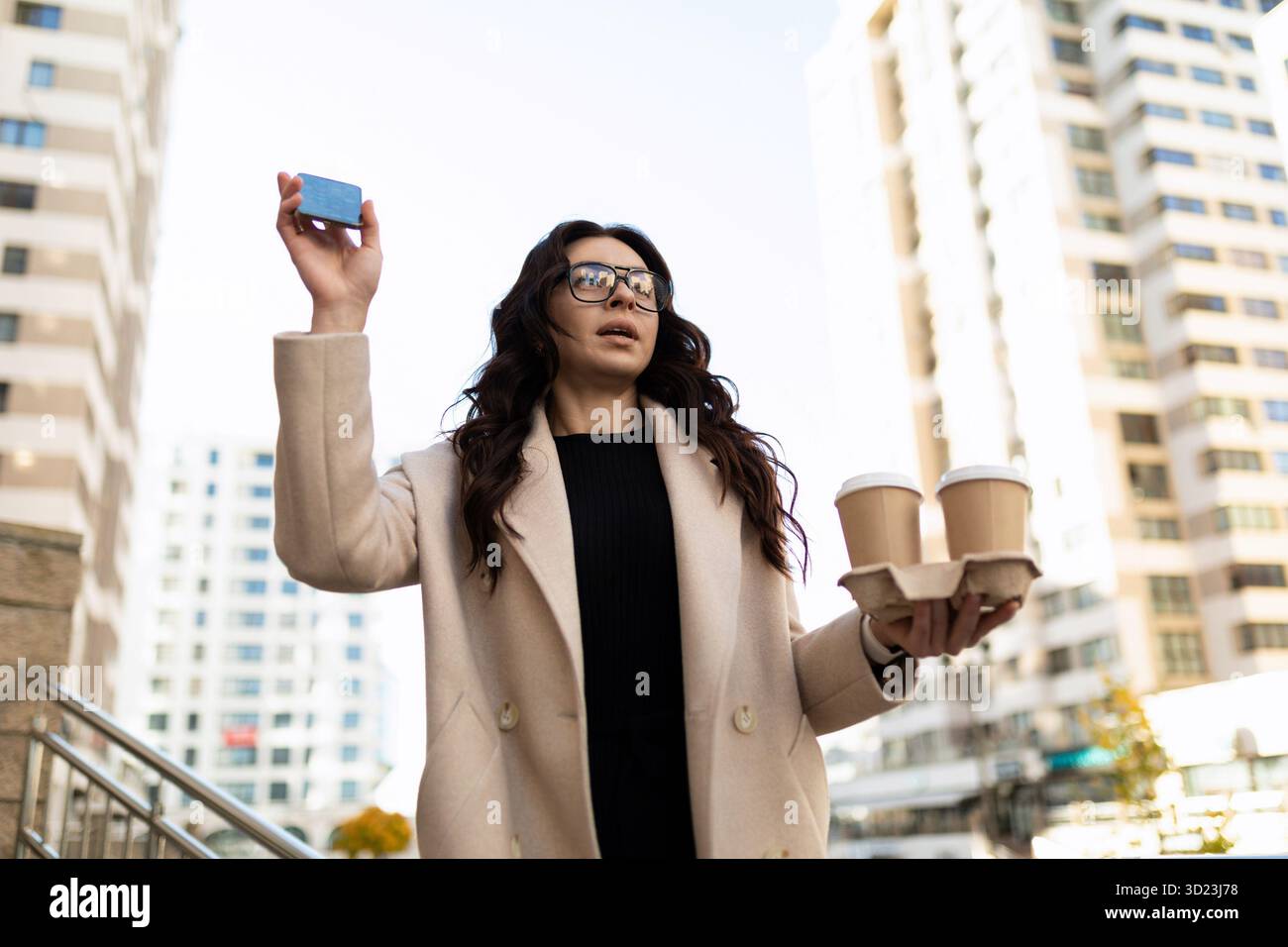 Junge, stilvolle Frau mit zwei Tassen Kaffee in den Händen, die einem Freund in der Stadt winkt Stockfoto