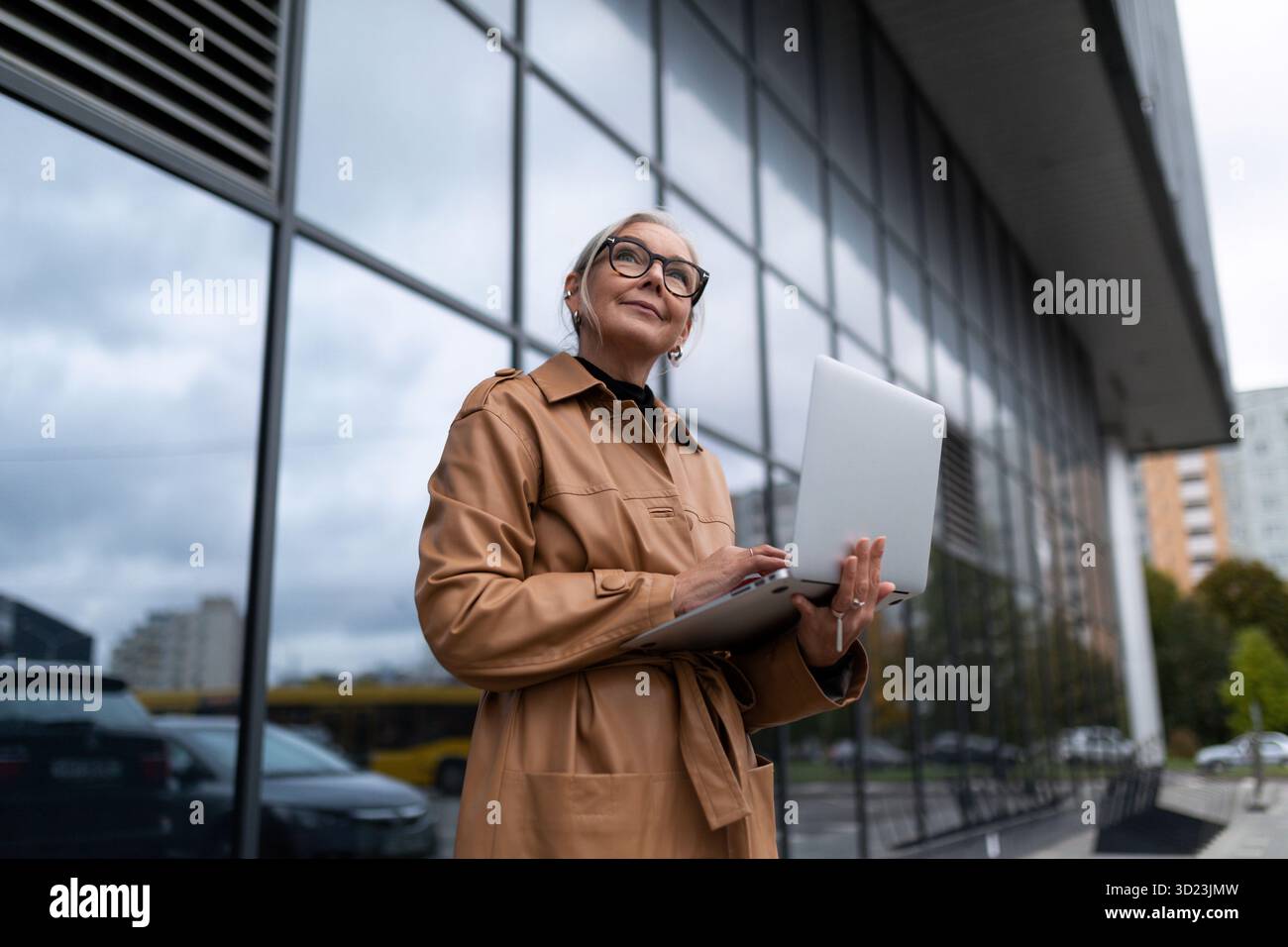 Geschäftsfrau in einem Ledermantel mit einem Laptop in der Hand vor dem Hintergrund einer Glasfassade des Büros Stockfoto