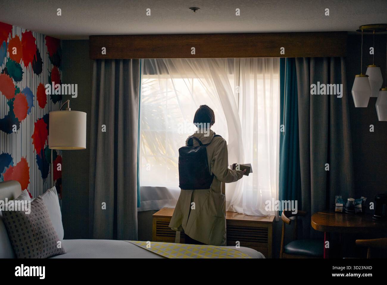 Frau mit Rucksack steht am Hotelfenster in einem stilvollen Hotelzimmer mit lebhafter Wanddekoration. Kalifornien, USA Stockfoto