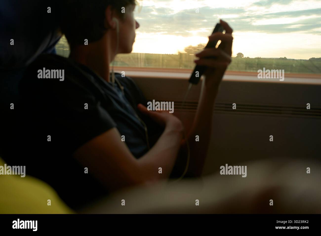 Junger Mann in einem Zug, der Kopfhörer trägt, aus dem Fenster auf Sonnenuntergang blickt und das Telefon hält. München, Deutschland Stockfoto