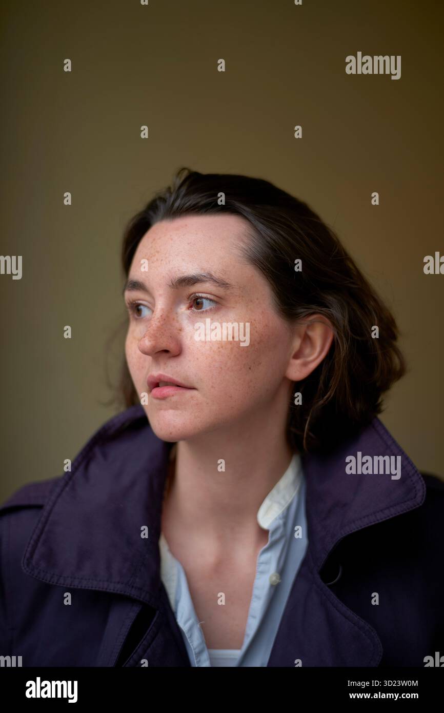 Nachdenkliche junge Frau in einem lila Mantel, die vor neutralem Hintergrund zur Seite blickt. Brüssel, Belgien Stockfoto