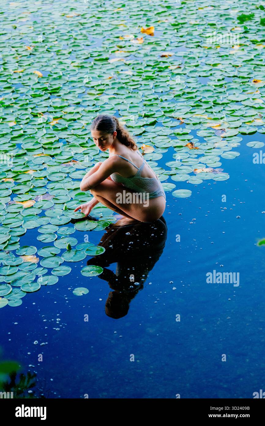 Frau, die sich auf Lilienpolstern über einer ruhigen blauen Wasseroberfläche hockt, in einer ruhigen Szene. Wildcat Lake Park, WA, USA Stockfoto
