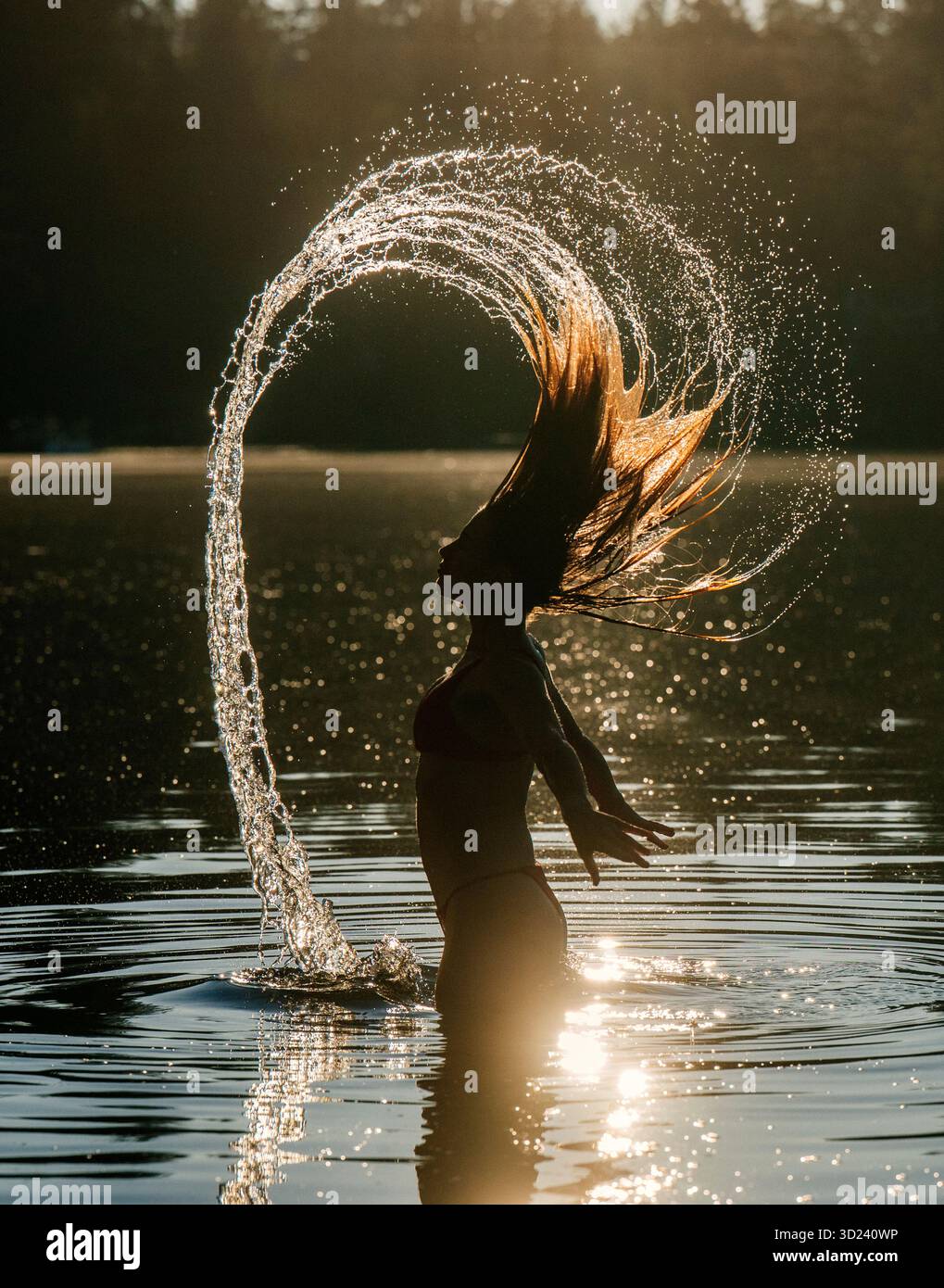 Frau, die bei Sonnenuntergang im Wasser Haare wirft, einen Tropfenbogen und eine ruhige Szene erzeugt. Wildcat Lake Park, WA, USA Stockfoto