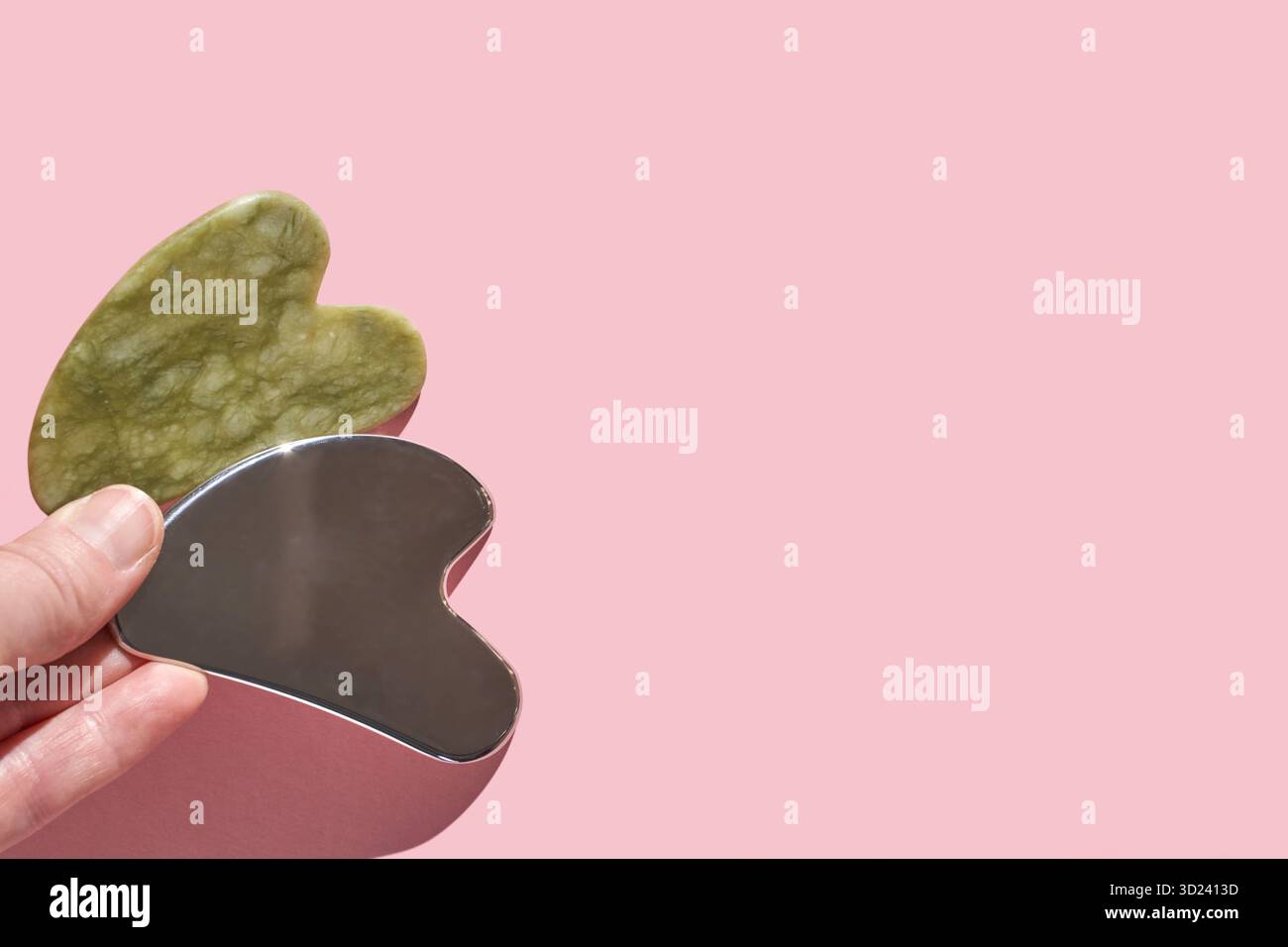 Gua-Sha-Werkzeuge: Grüne Jade und Edelstahl-Massagestein auf zartem Rosa Stockfoto