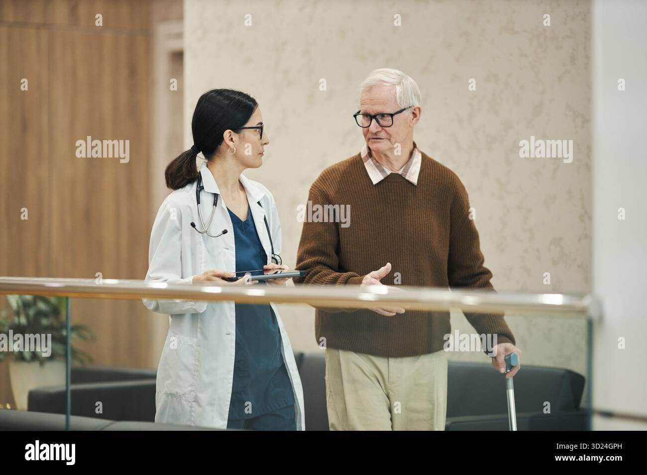 Asiatische Frau mittleren Alters Doktor geht und spricht mit älteren kaukasischen Mann, der Stock in der medizinischen Einrichtung hält, beide im Gespräch, Doktor hält digitale Tablette Stockfoto