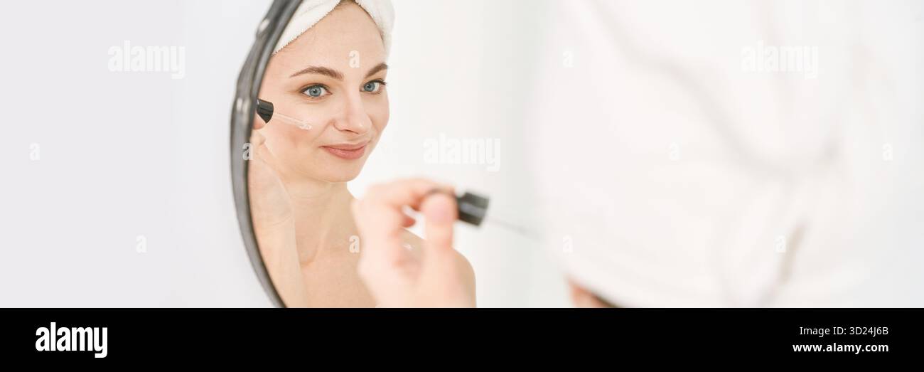 Junge kaukasische Frau, die Hautpflegeserum im Bad mit Handtuch umwickelt aufträgt Stockfoto