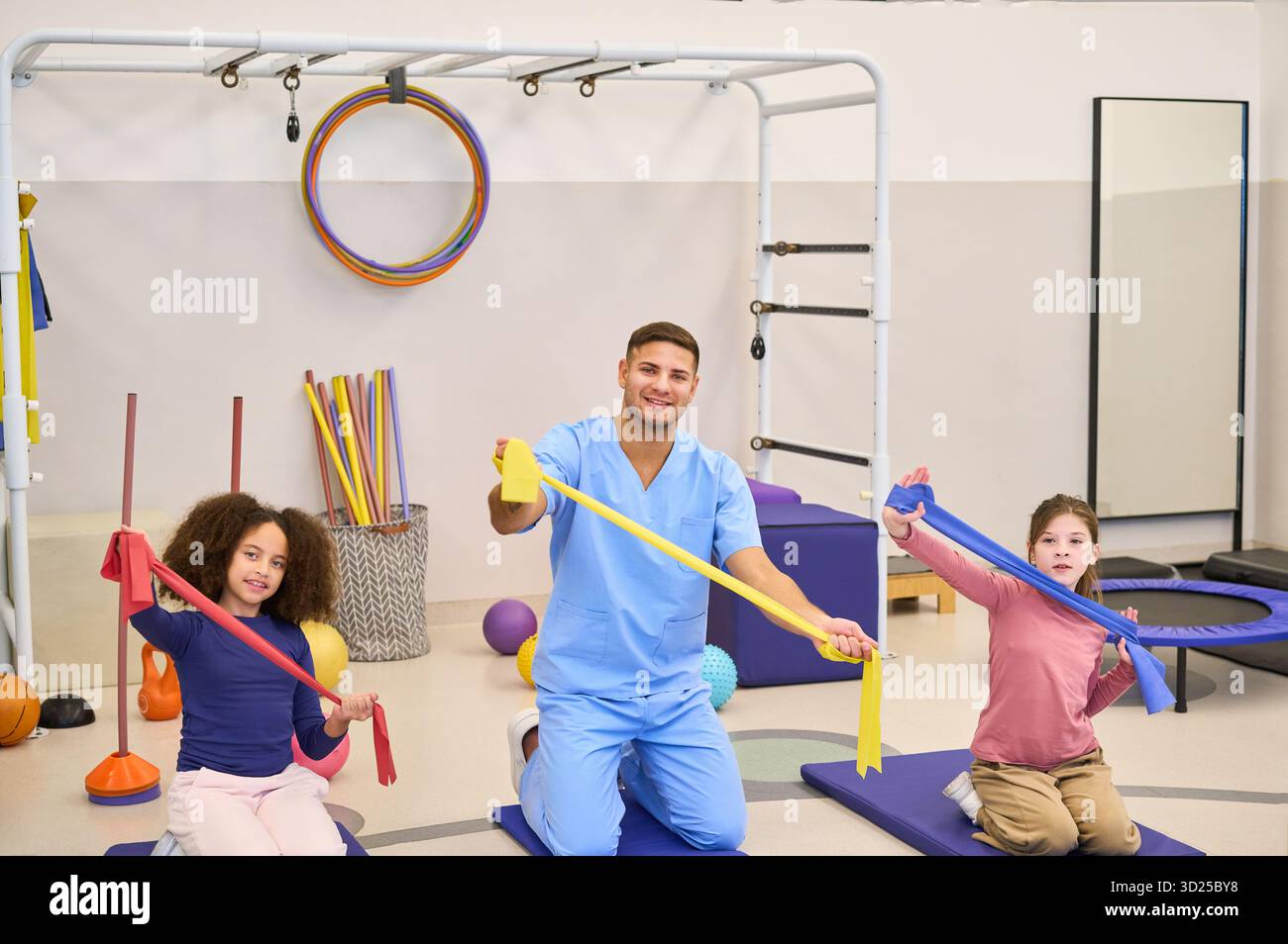 Ein Therapeut führt zwei Kinder in einer spielerischen Reha-Übung mit bunten Bändern in einer pädiatrischen Physiotherapie-Umgebung. Die Sitzung konzentriert sich auf Motor Stockfoto