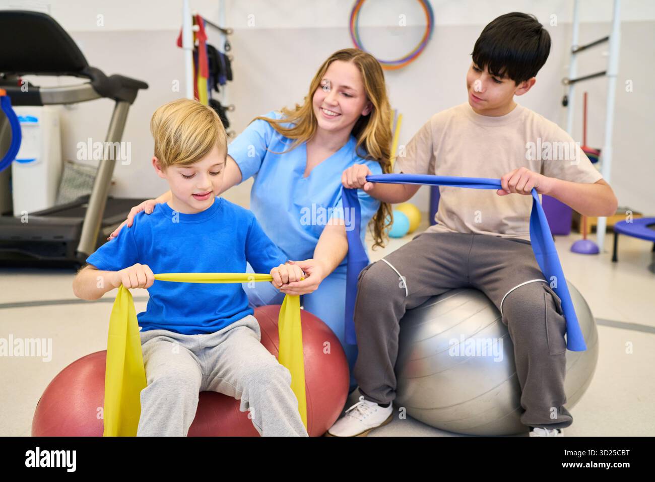 Ein freundlicher Therapeut hilft zwei Kindern während einer Rehabilitationssitzung, indem er Widerstandsbänder und Stabilitätsbälle verwendet, um Gleichgewicht, Kraft und Moto aufzubauen Stockfoto