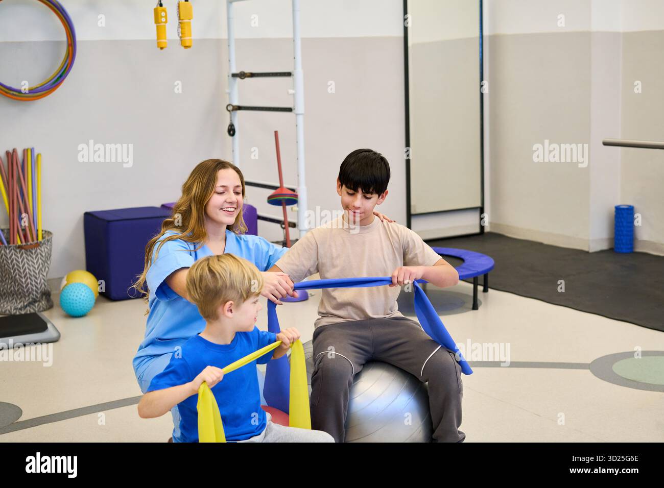 Ein pflegender Therapeut arbeitet mit zwei Kindern in einer pädiatrischen Reha-Umgebung, verwendet Widerstandsbänder und einen Stabilitätsball, um motorische Fähigkeiten und Balance zu üben Stockfoto