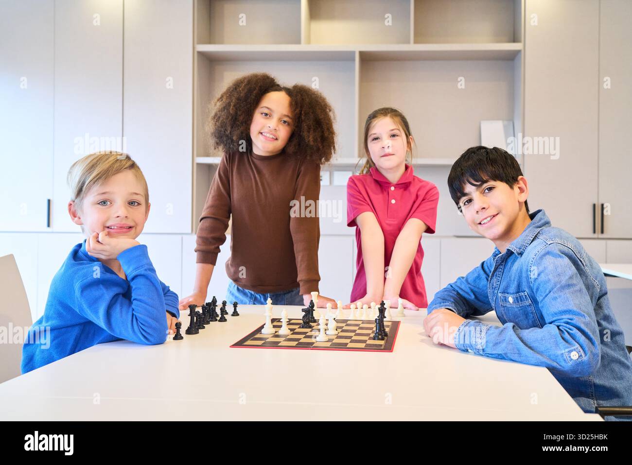 Eine Gruppe von Kindern sitzt und steht um ein Schachbrett in einem hellen, modernen Raum. Sie lächeln und nehmen an einer sozialen, pädagogischen Aktivität teil, die sie fördern Stockfoto