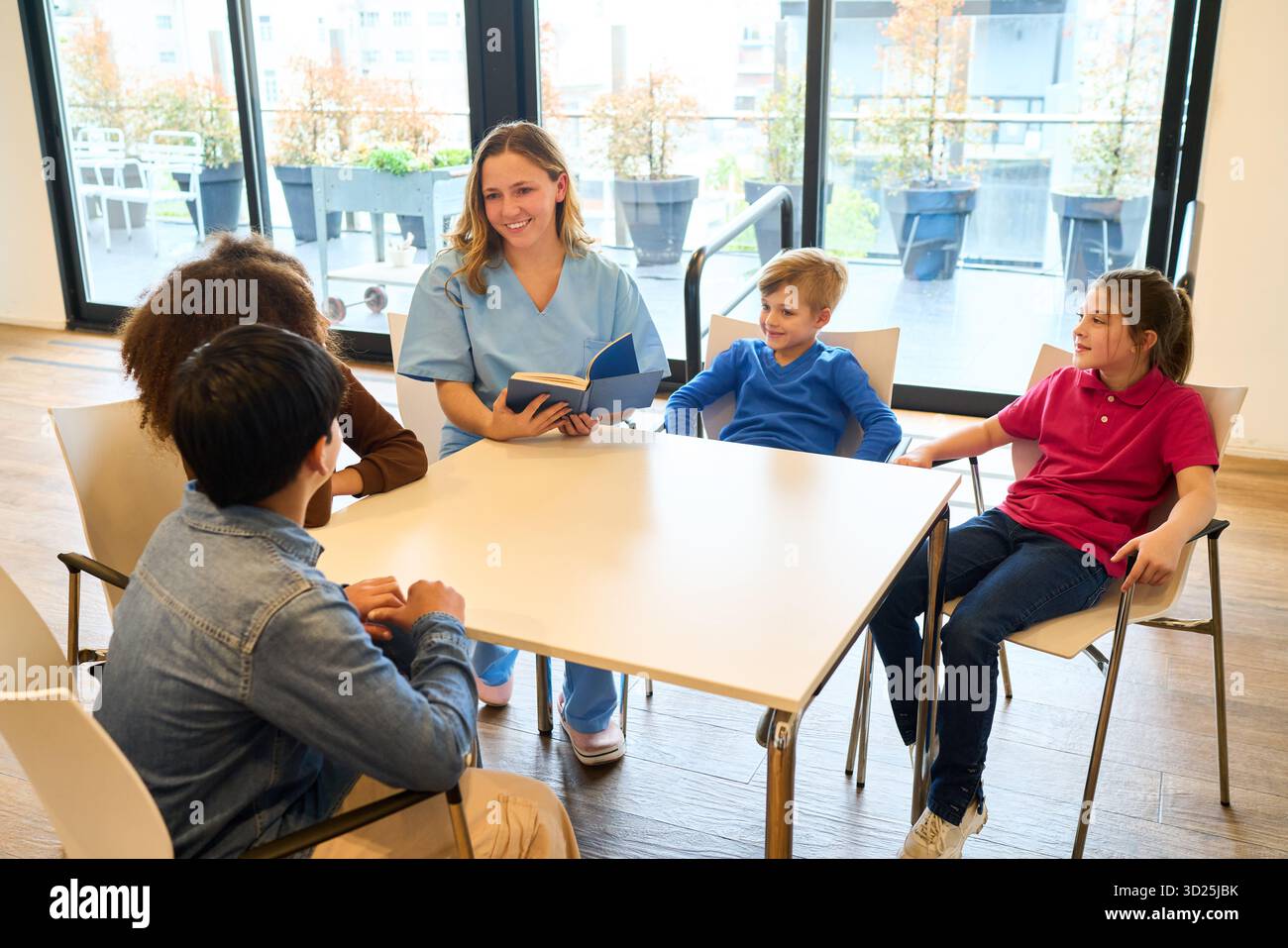 Ein freundlicher Therapeut führt eine kleine Gruppe von Kindern in einer Reha-Aktivität um einen Tisch, um die Motorik, den Fokus und die soziale Hilfe zu fördern Stockfoto