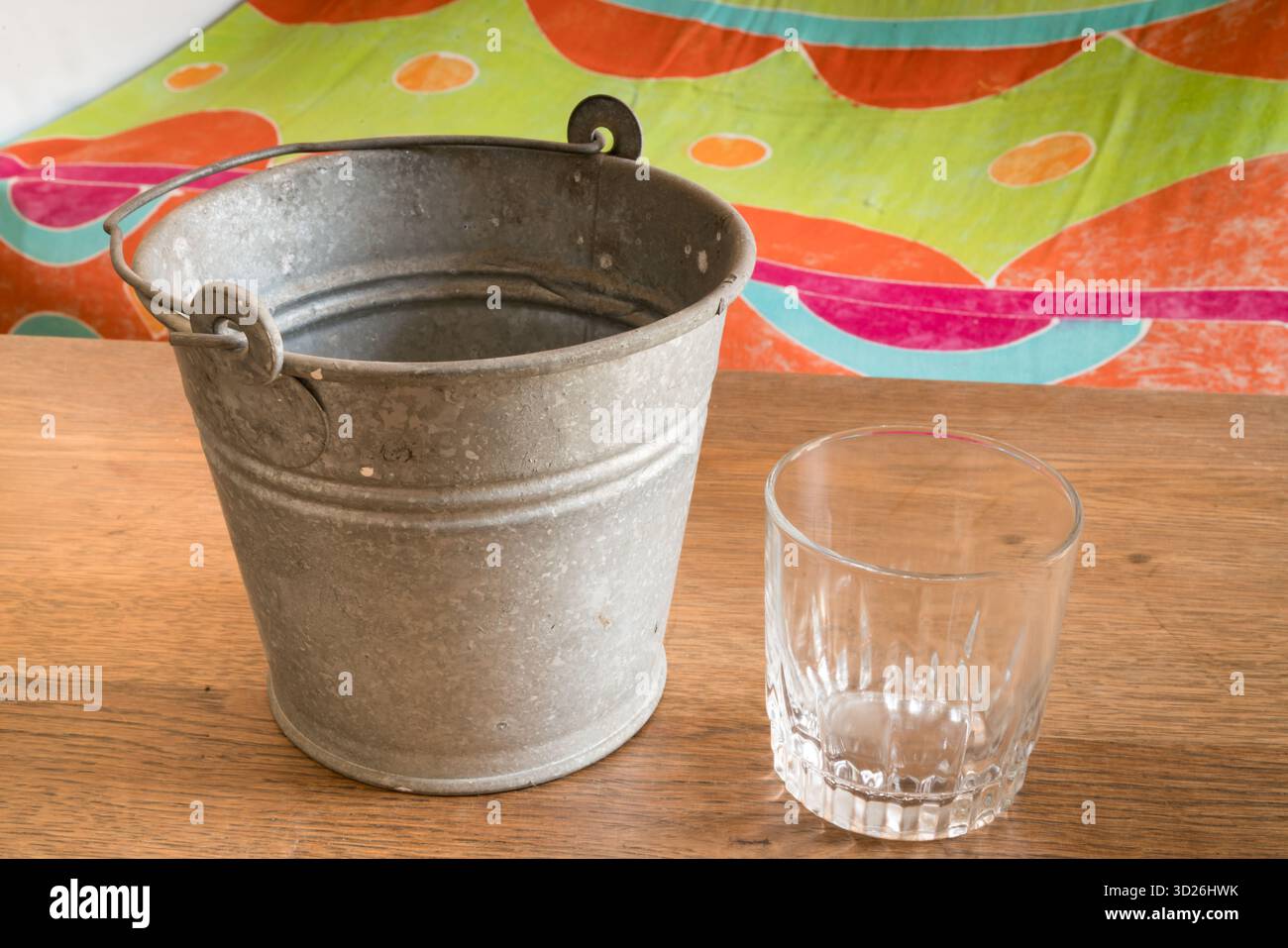 Leeres Glas und verzinkter Metalleimer auf Holzoberfläche mit farbenfrohem Hintergrund Stockfoto