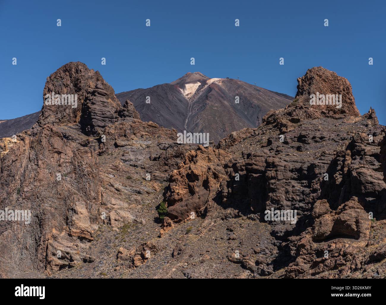 Zerklüftete rötliche vulkanische Felsformationen ragen vor dem Teide unter klarem blauen Himmel in den Vordergrund Stockfoto