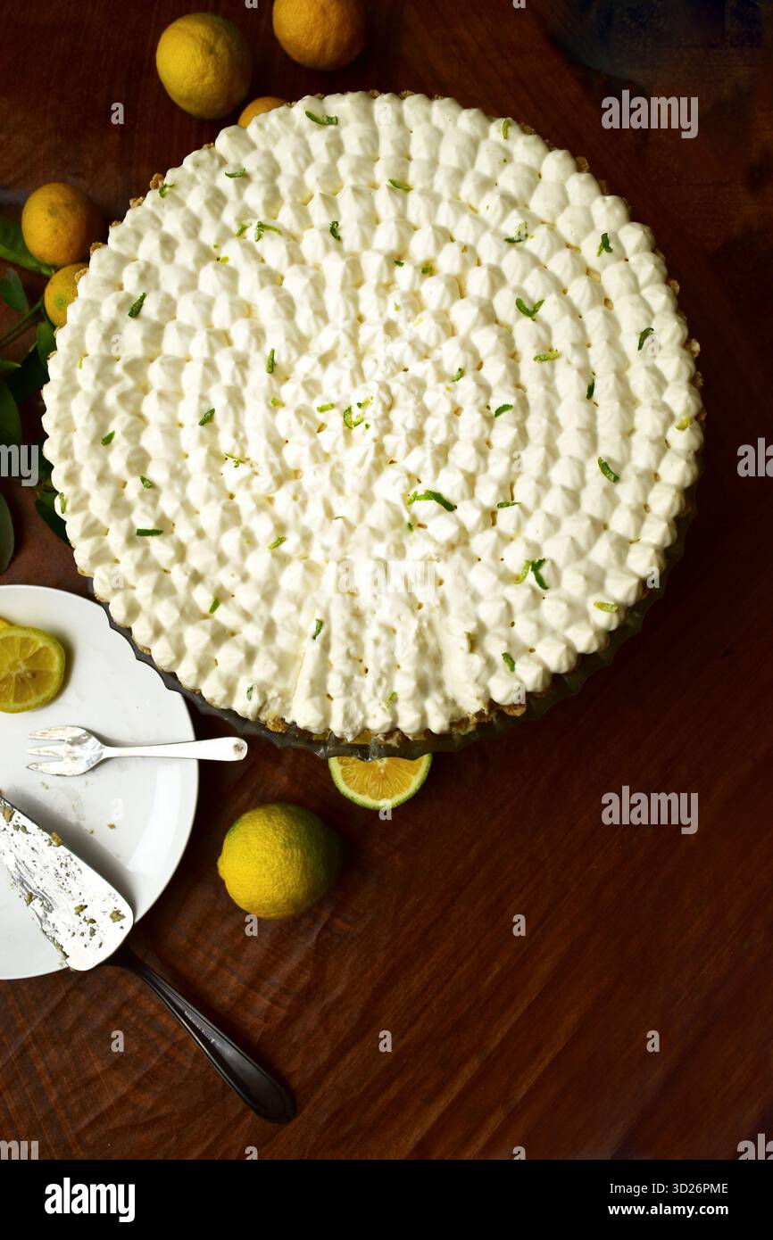 Hausgemachte Zitronenkuchen auf einem Glaskuchenständer, dekoriert mit Schlagsahne und Limettenschale. Ein Stück des cremigen Desserts wird auf einem weißen Teller serviert Stockfoto