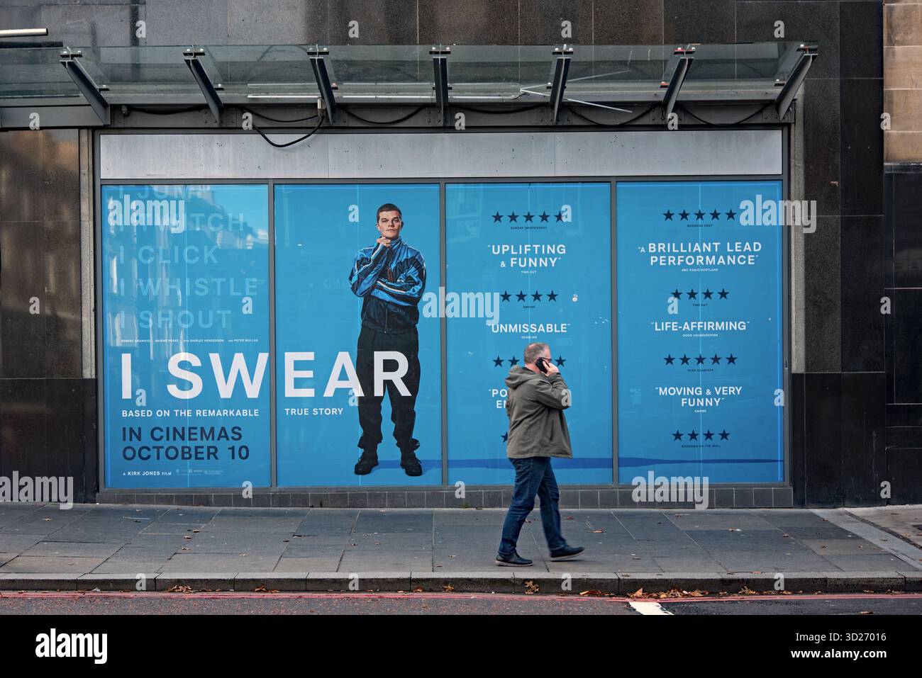 Mann läuft mit Telefon vorbei an der Werbung für den Film „I Swear“ in Edinburgh, Schottland, Großbritannien. Stockfoto
