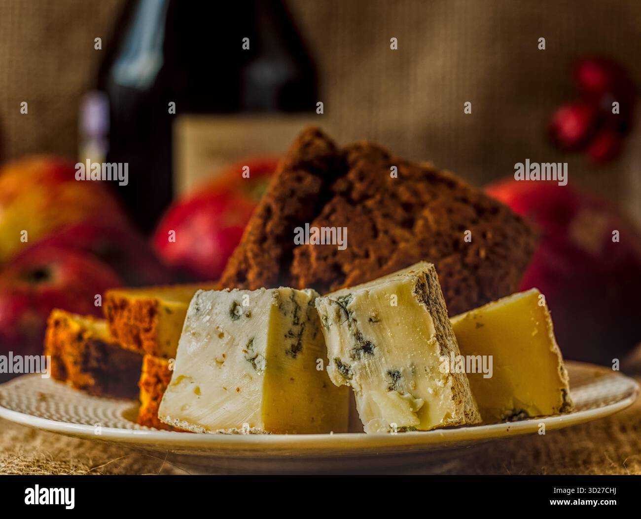 Italienischer Blauschimmelkäse Gorgonzola und Pfeffer Jack Käse auf Teller, schwarzer Brandy oder Weinflasche und rote Äpfel dahinter, dunkle gemütliche Atmosphäre Stockfoto