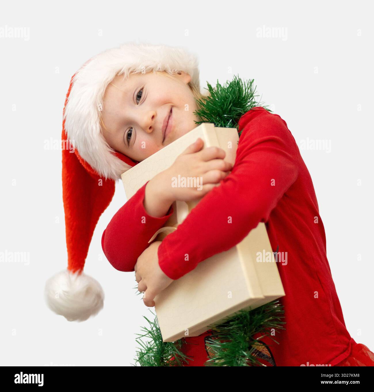 Ein fröhliches Kind in rotem Hemd und Weihnachtsmann-Hut umhüllt ein Geschenk, das während der Weihnachtszeit zu Hause Spannung und Glück ausstrahlt. Stockfoto