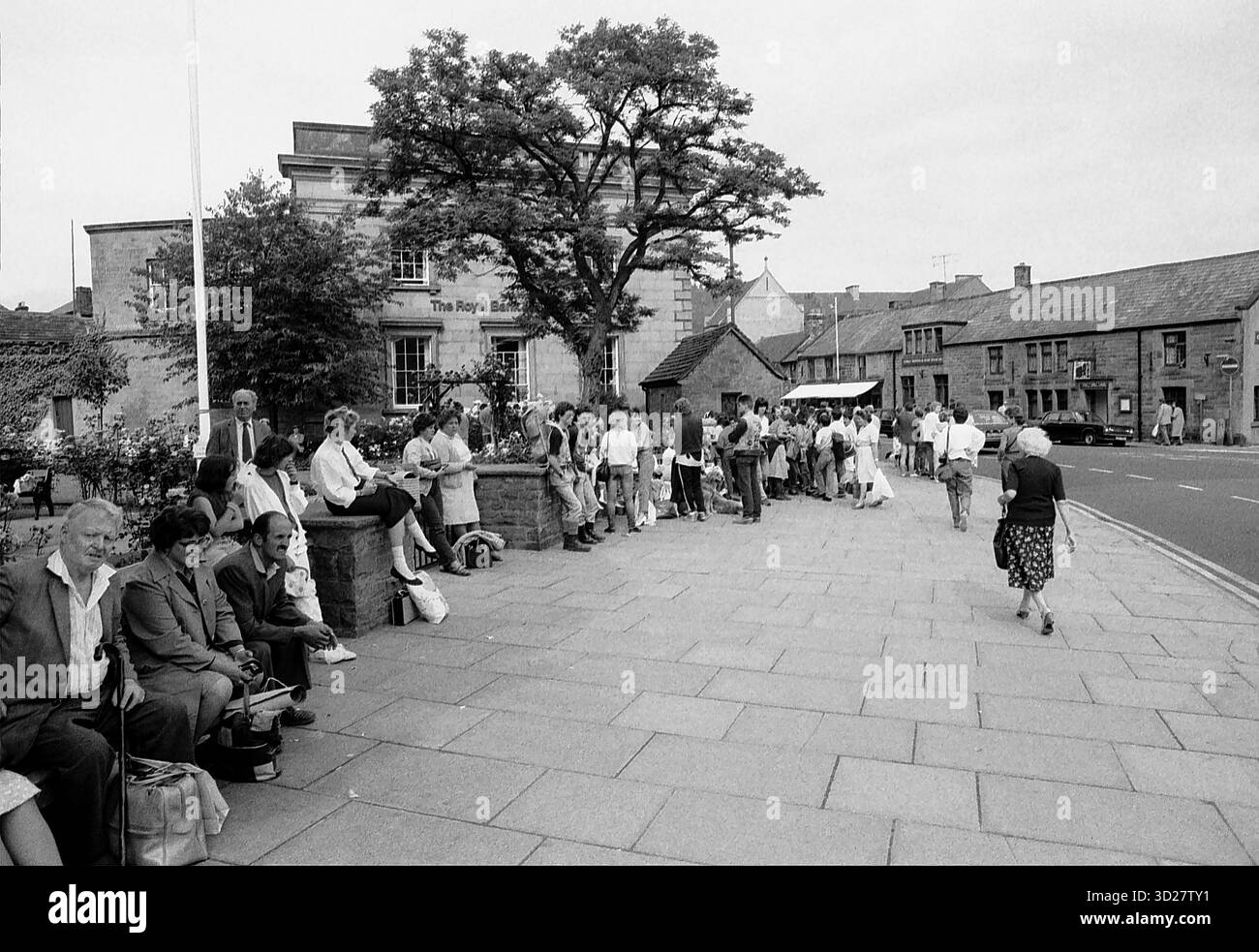 Ein Schwarzweißfoto fängt eine geschäftige Szene in einer kleinen englischen Stadt ein. Eine Gruppe von Fußgängern, vor allem Erwachsene und Kinder, ist auf einem gepflasterten Platz versammelt und wartet auf einen Bus. Die Szene wird von einem prächtigen Gebäude, „The Royal Oak“, und alten Bäumen dominiert, die einen Einblick in das tägliche Leben und den architektonischen Charme einer historischen Stadt in Großbritannien bieten. Stockfoto