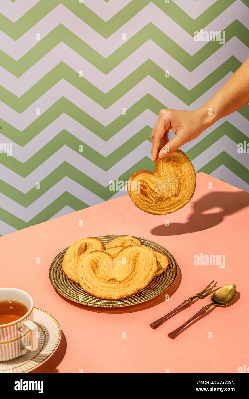 Herzförmige Palmier Cookies mit Hand heben eins auf rosa Tisch und Teetasse im Chevron Hintergrund Stockfoto