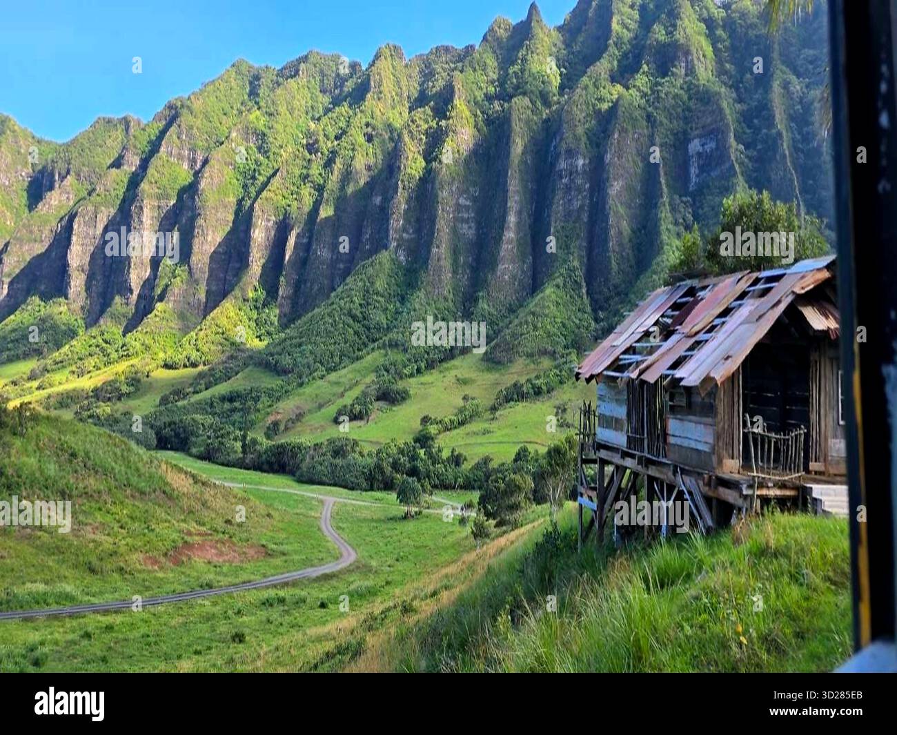 Rustikale Holzhütten überblicken das üppige Kualoa Ranch Tal auf Oahu, Hawaii, mit dramatischen grünen Ko'olau Bergkämmen und einem gewundenen Schotterpfad. Stockfoto