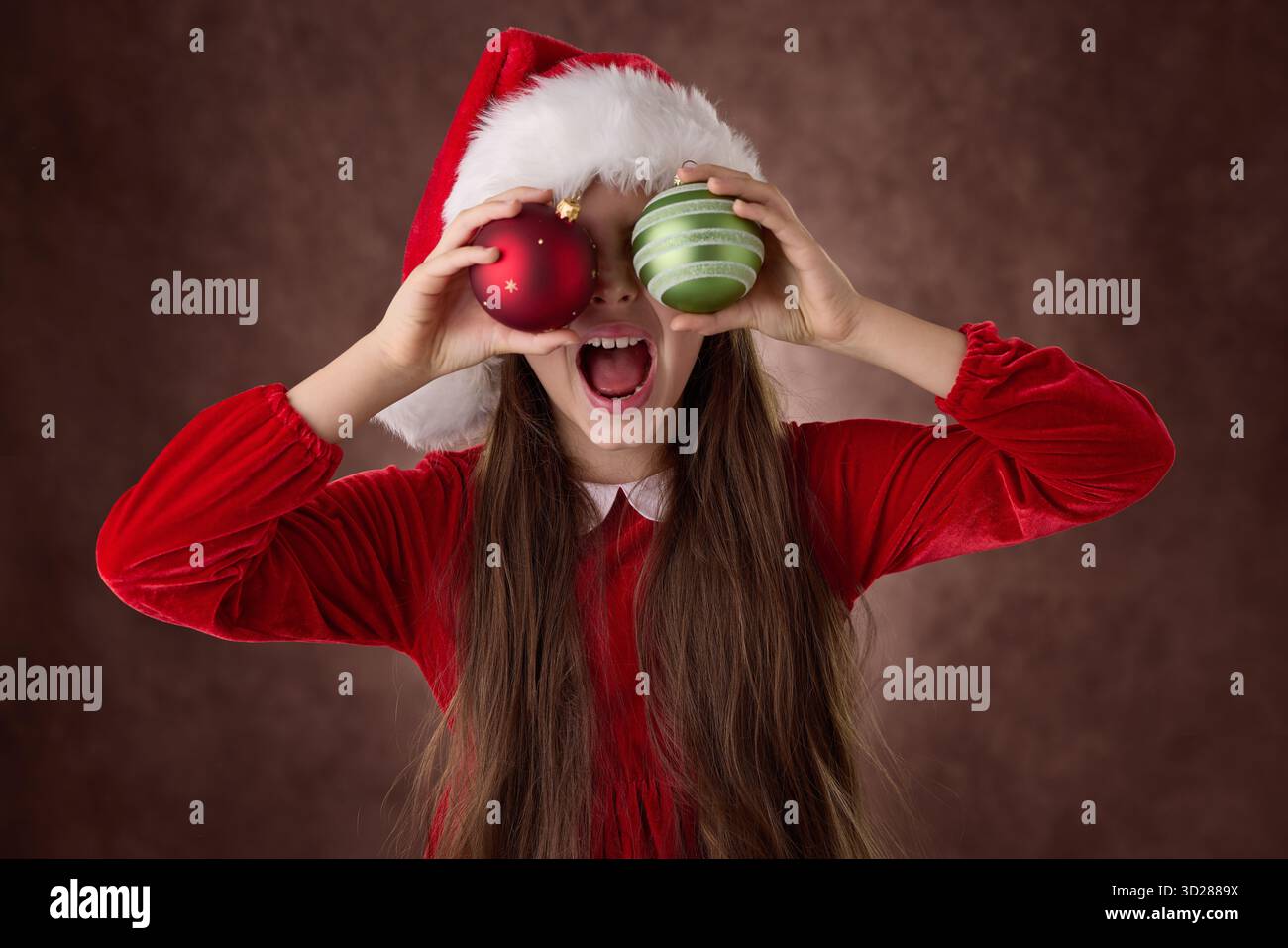 Lächelndes Mädchen hält Weihnachtsbaumkugeln in seinen Augen und trägt Santa Claus Hut Kleid Porträt im Vintage Hintergrund Stockfoto