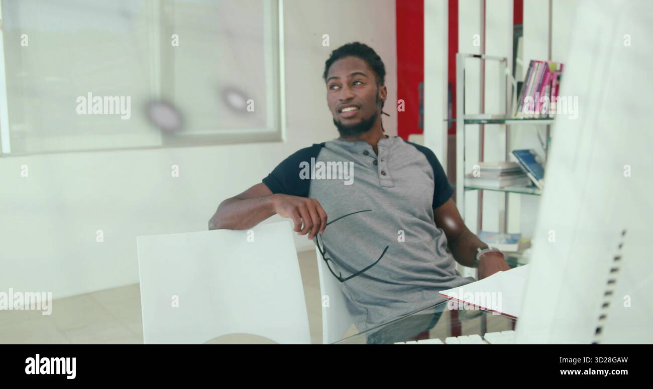 Lehnender Mann, der eine Brille hält, auf den Glasschreibtisch im Büro blickt, mit Monitor, Büchern Stockfoto