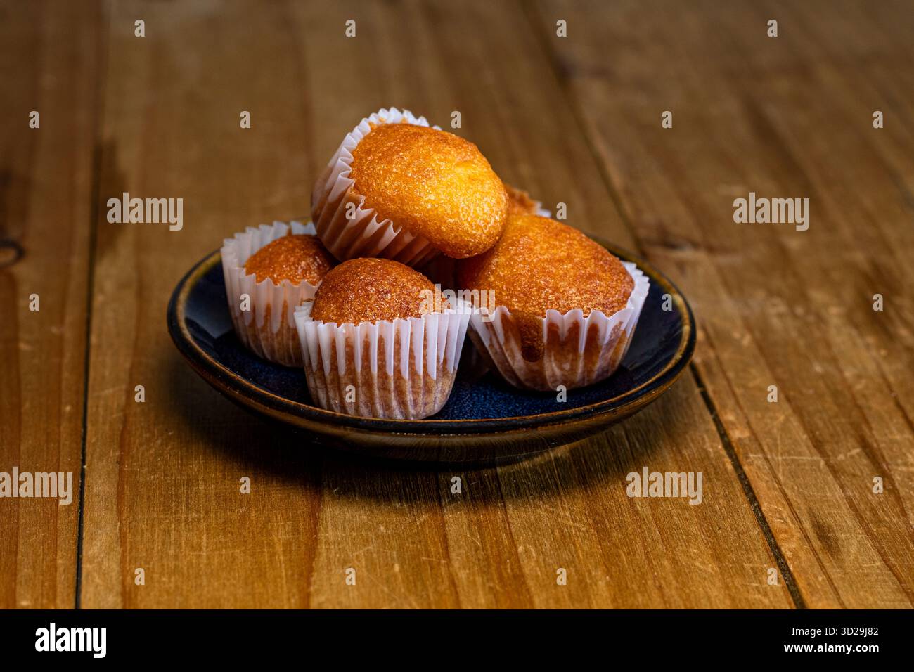 Goldene gebackene Mini-Muffins in Pappbechern, präsentiert auf einem dunklen Teller, auf einem strukturierten Holztisch Stockfoto