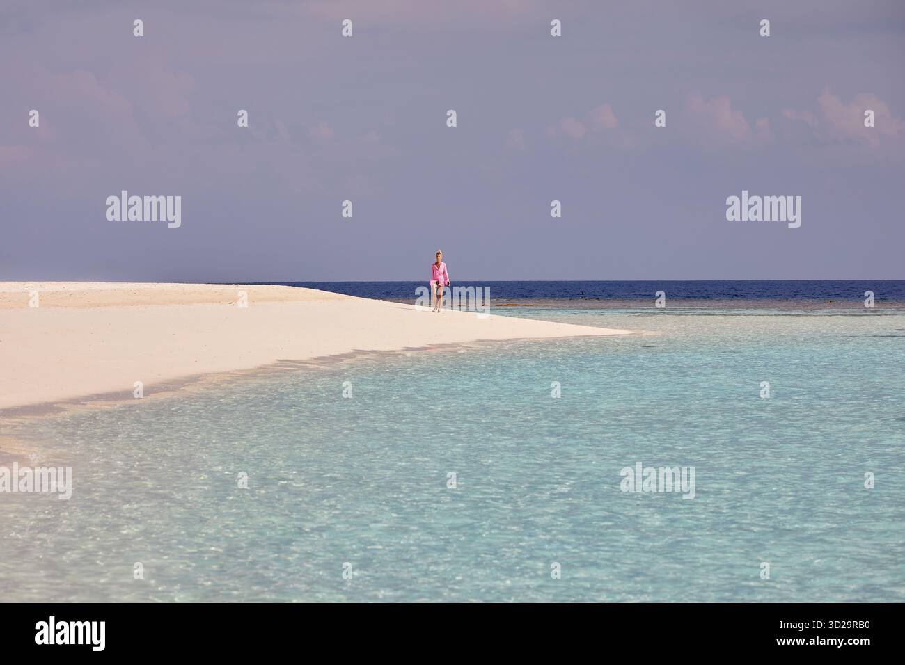 Eine Frau, die allein in hellem Sonnenlicht an einem tropischen weißen Sandstrand auf den Malediven spaziert, umspült von ruhigem aquamarinem Wasser. Stockfoto