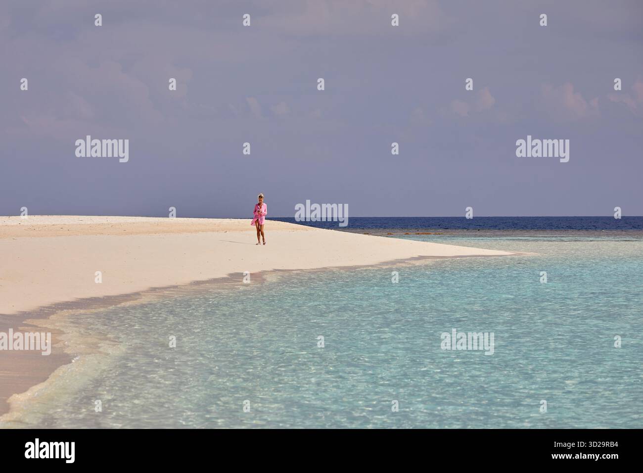 Eine Frau, die allein in hellem Sonnenlicht an einem tropischen weißen Sandstrand auf den Malediven spaziert, umspült von ruhigem aquamarinem Wasser. Stockfoto