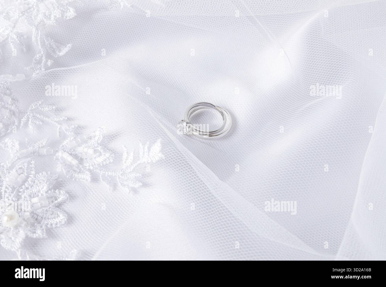 Zwei Eheringe aus weißem Gold mit einem Diamanten liegen auf einem weißen Schleier mit weichen Falten. Hochzeitshintergrund. Layout für Design. Postkarte. Einladung Stockfoto