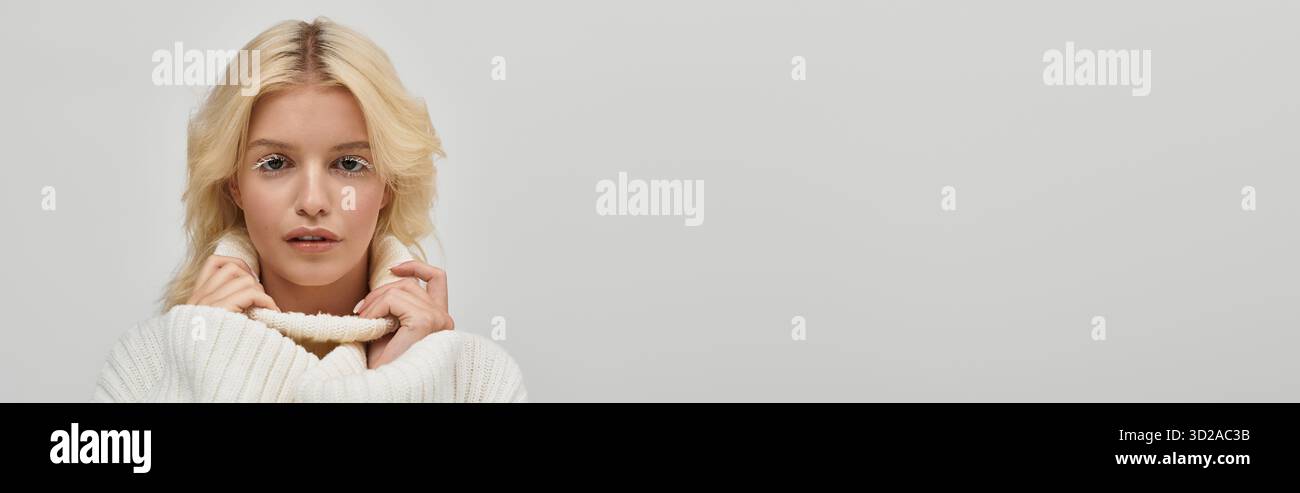 Eine junge blonde Frau mit atemberaubenden weißen Wimpern posiert warm in einem gemütlichen Strickpullover, Banner Stockfoto