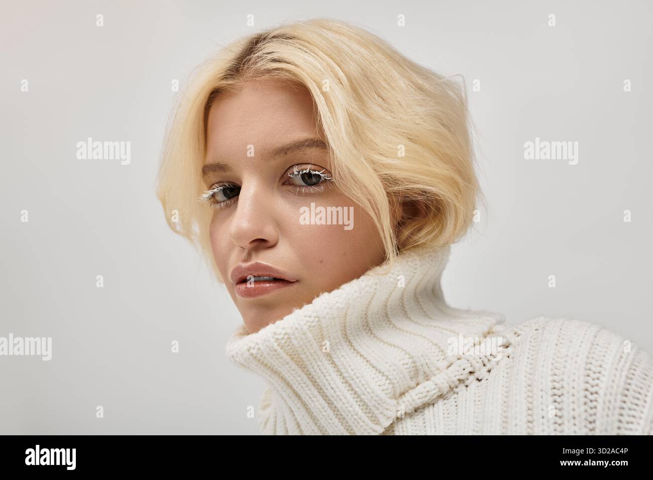 Eine junge blonde Frau mit weißen Wimpern posiert anmutig in einem gemütlichen Strickpullover. Stockfoto