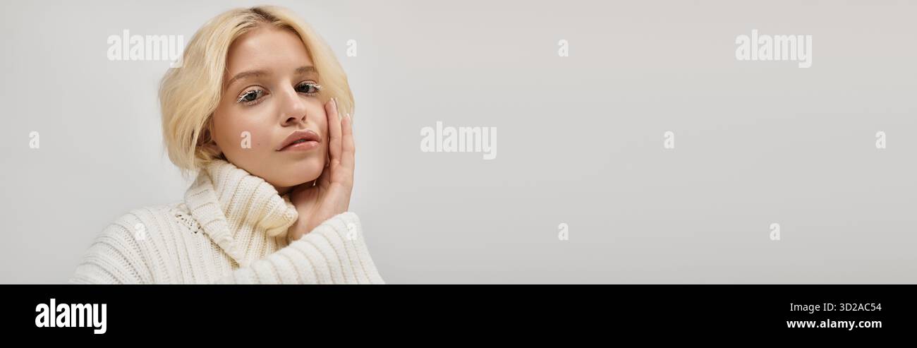 Eine junge blonde Frau mit weißen Wimpern legt ihre Hand auf ihrem Gesicht in einem gemütlichen Pullover, Banner Stockfoto
